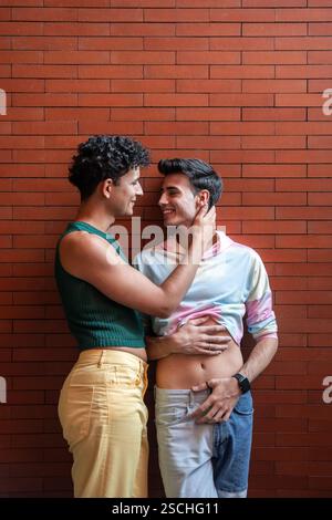 A joyful gay couple embracing and smiling at each other, standing against a red brick wall. The tender moment captures love and connection in an urban Stock Photo