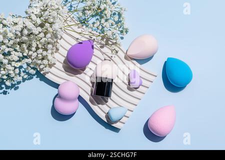 From above shot of various colorful makeup sponges, including beauty blenders, displayed elegantly on a textured mat alongside white blossoms on a pal Stock Photo