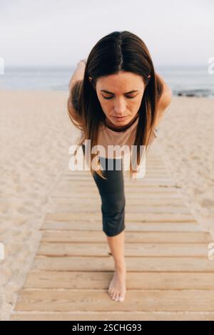 Focused woman with eyes closed practicing an advanced yoga pose on a wooden path at the beach. She is focused on balancing, enhancing her strength and Stock Photo