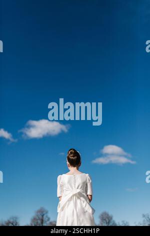 A girl in a white First Communion dress is seen from behind, standing under a bright blue sky with soft clouds. Her hair is neatly styled, symbolizing Stock Photo