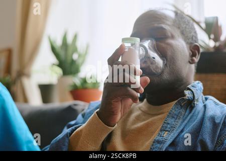 Low angle shot of senior African American man suffering from asthma sitting on sofa and using nebulizer Stock Photo