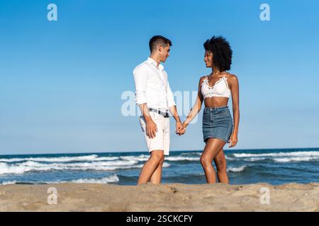 A young interracial couple enjoying a romantic walk on the beach, holding hands and smiling under the summer sun. Stock Photo