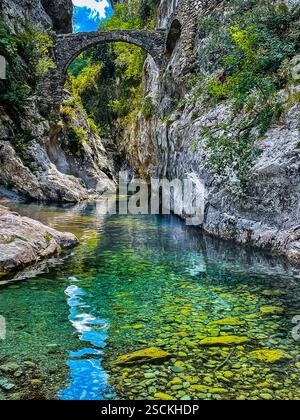 Calm Stream with Stone Bridge in a Natural Landscape A tranquil stream with turquoise-green water flowing under an ancient stone bridge, surrounded by Stock Photo