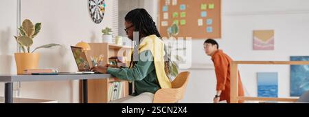 Woman with long braids working on laptop in modern office space with green plants on table. Colleague walking in background Stock Photo