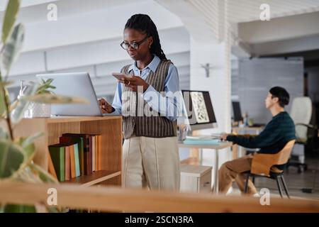 Young professional standing in modern office analyzing data on smartphone while using laptop. Other employees working in background creating dynamic work environment Stock Photo