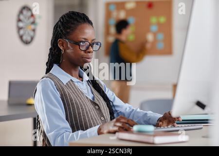 Black woman wearing glasses concentrating while working on laptop at office desk in open office layout with colleagues collaborating in background Stock Photo