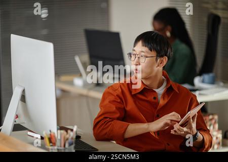 Asian employee working diligently on a tablet in a contemporary open-plan office with computers, while colleagues are in background, focusing on tasks Stock Photo