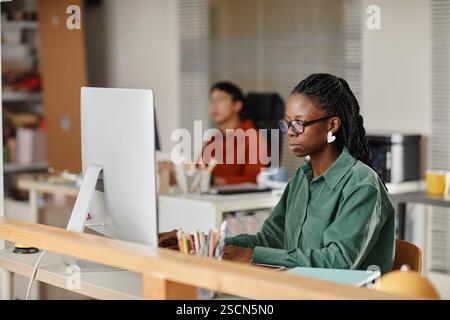 Young professional focused on computer screen in modern office environment showing serious expression. Colleagues and office supplies are visible in background Stock Photo