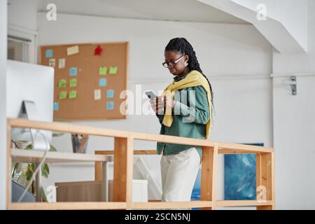 African American woman wearing glasses, using smartphone in contemporary office space with bulletin board behind her, standing near wooden railings Stock Photo