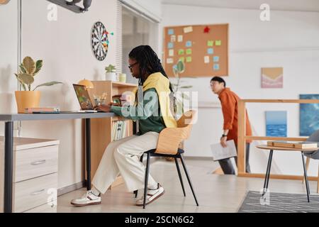 Two professionals working in a modern office setting, with one seated at desk using laptop and another walking in background holding documents Stock Photo