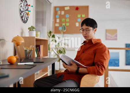 Young man immersed in reading while seated at modern wooden desk with organized bookshelves and open laptop in cozy home office setting Stock Photo