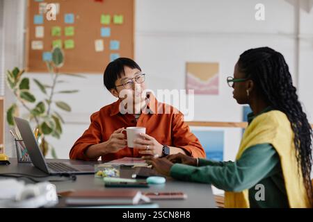 Asian man and Black woman engaging in conversation in modern office environment with notes on wall. Both seated at desk with plants in background, exchanging focused ideas Stock Photo