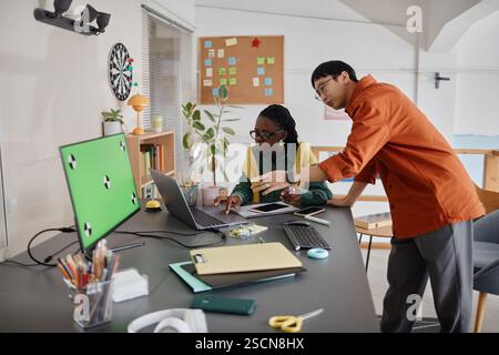 Two professionals working together at a desk with a laptop and various office supplies, discussing ideas and sharing insights into their project planning Stock Photo