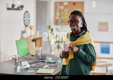 Smiling businesswoman standing in office holding coffee cup while looking relaxed and happy. Desk with computer and office supplies in background, creating productive atmosphere Stock Photo