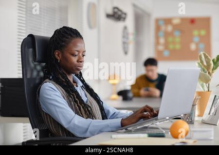 Woman sitting at desk in modern office, working on laptop and notepad, with colleagues in the background, focused on task Stock Photo