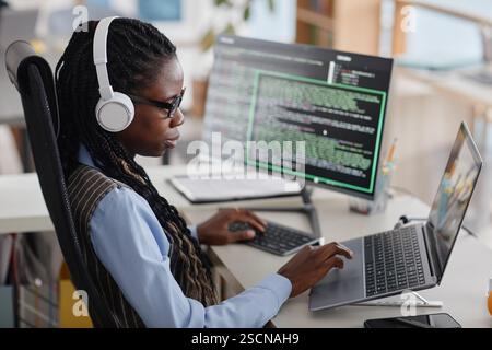 Female programmer deeply engaged in coding while wearing headphones and working with multiple screens in modern office environment Stock Photo