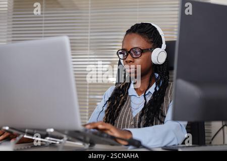 Portrait of woman wearing headphones and working on computer in an office environment. She has braids and is looking focused on her task Stock Photo
