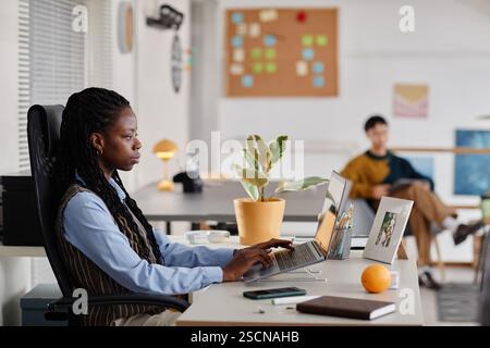 Black woman typing on laptop, working in modern office with colleagues and potted plants on desk Photo showing professional setting with collaborative and productive atmosphere Stock Photo