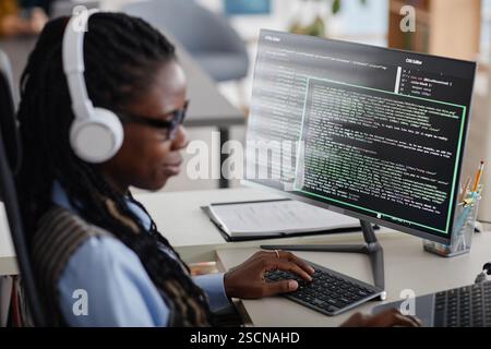 African American woman wearing headphones and working on computer programming in an office environment. She is deeply focused on coding displayed on a monitor Stock Photo