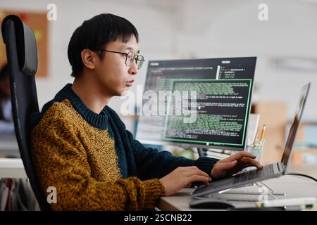 Man concentrating on writing code at workstation with multiple screens displaying programming codes. Wearing glasses and casual attire in modern office environment Stock Photo