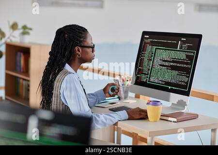 Woman engrossed in computer coding, sitting in modern office, using iMac and handling mouse. Office space featuring bookshelves in background, coffee cup, and nearby headphones Stock Photo
