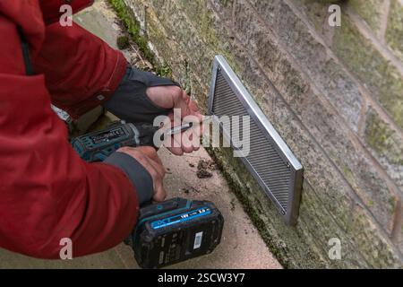 A man wearing a red jacket and fingerless gloves working outside. Man using power drill to screw a mesh cover onto a wall over an air brick. Stock Photo