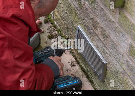 A man wearing a red jacket using an electric drill to screw a mesh cover onto a wall. The cover is over an air brick to prevent mice from entering. Stock Photo