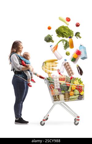 Mother with a baby in a carrier and a shopping cart full of falling food assortment isolated on white background Stock Photo