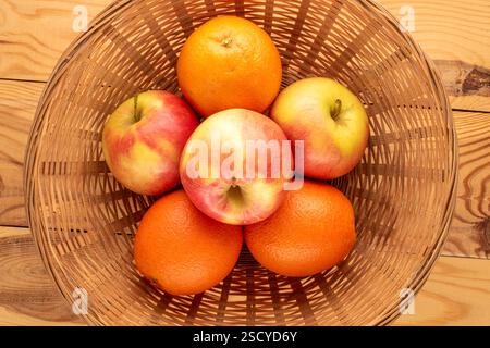 Three juicy oranges and three apples in a straw plate on a wooden table, close-up, top view. Stock Photo