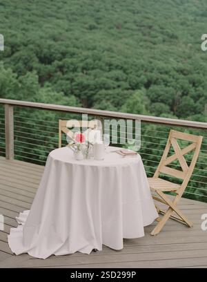 A beautifully set table with a white tablecloth and floral arrangement beside an empty chair on a wooden deck. Stock Photo