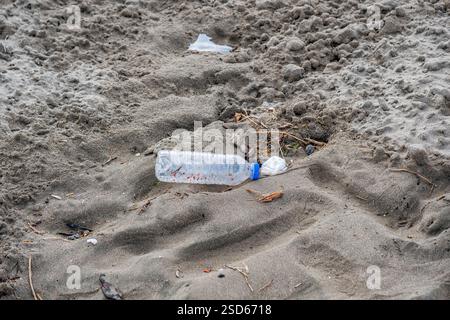 A bottle of water is laying on the sand. The bottle is empty and has a blue cap. The bottle is surrounded by sand and debris, indicating that it has b Stock Photo