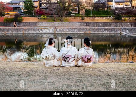 Three young women sit wearing traditional clothes on the banks of the Kamogawa (Kamo River), the main waterway running through Kyoto City, Japan. Stock Photo
