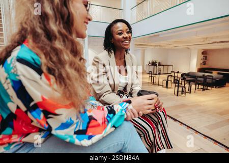Two colleagues are sitting in a contemporary coworking office, engaging in an informal chat while enjoying coffee. The setting conveys a collaborative Stock Photo