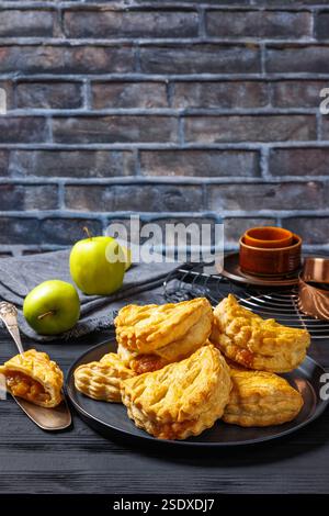 puff pastry apple turnovers on a plate on black wooden table with plates, napkin, apples, cake shovel and brick wall in background, vertical view from Stock Photo
