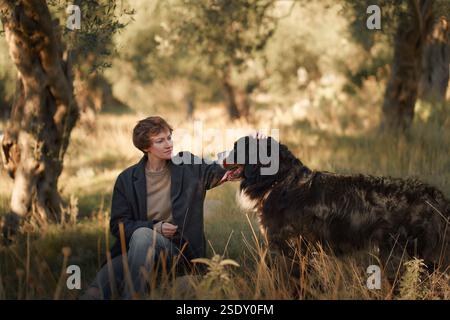 A woman sits with her Bernese Mountain Dog in a grassy area, enjoying the peaceful surroundings. Stock Photo