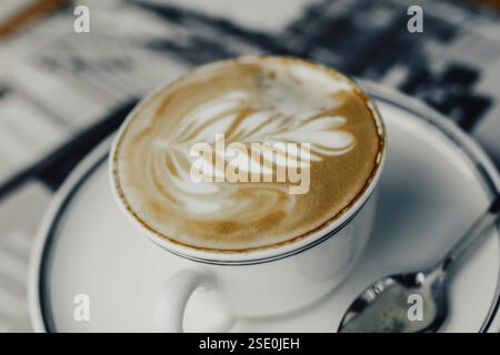 Coffee cup with leaf pattern latte art, accompanied by a spoon on a saucer Stock Photo