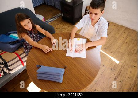 Two children sit at a round wooden table, collaborating on an educational task, inside a cozy home setting. Stock Photo