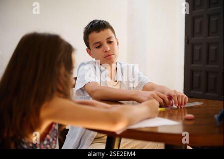 Two children are sitting at a table, appearing focused as they collaborate on a shared task, likely involving writing or crafting. Stock Photo