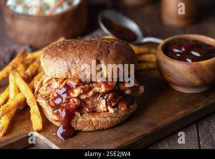 Barbeque pulled chicken sandwich on whole wheat bun with french fries on wooden cutting board and coleslaw in background Stock Photo