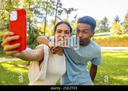 Multi-ethnic couple taking selfie gesturing victory and blowing a kiss in a park Stock Photo