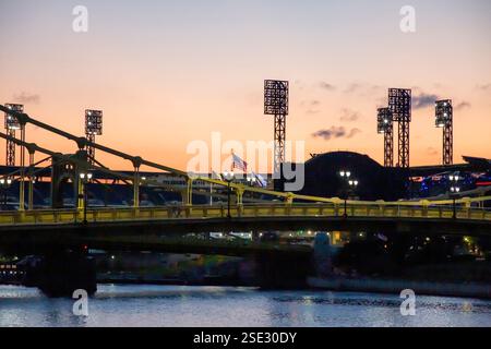 The view shows a vibrant sunset illuminating a bridge while stadium lights twinkle in the background. Stock Photo