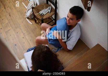 Two children engage in conversation while sitting on stairs, surrounded by school supplies. They appear to be on a break or studying together, demonst Stock Photo