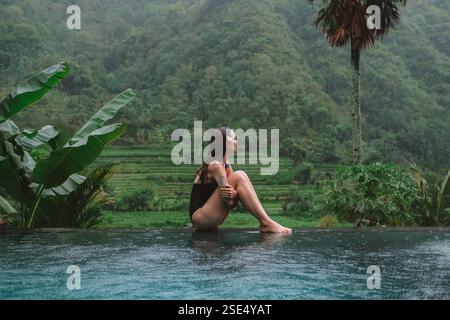 Young woman in a swimsuit sits by an infinity pool surrounded by tropical plants Stock Photo