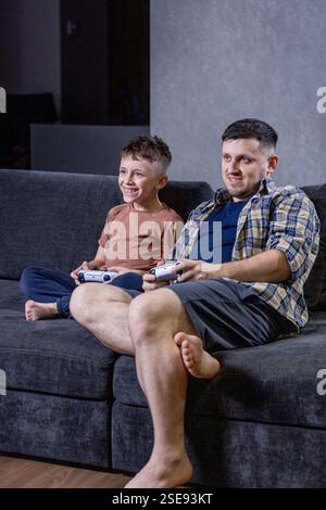 Father and his little son sit on sofa in living room playing video games together at home. Stock Photo