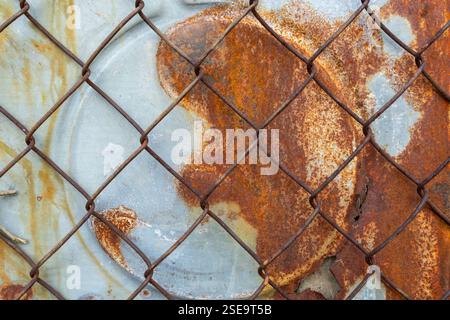A close-up view of a rusty metal surface partially obscured by a chain-link fence. The textures showcase orange and brown rust with a weathered blue b Stock Photo