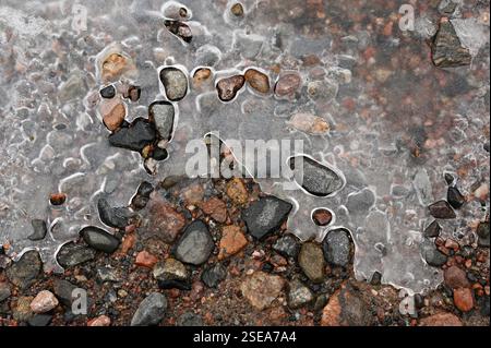 A close-up view of ice partially covering a rocky ground, capturing the intricate patterns formed as the ice begins to melt. The frozen surface reveal Stock Photo