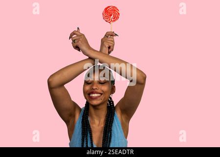 Happy young woman with braids holding red lollipop with crossed arms, smiling and standing on pink background Stock Photo