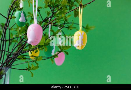 Colorful Easter eggs with cute white bunnies hanging on green tree branches, green background. Easter concept. Easter still life. Stock Photo