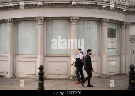 Pedestrians walk past a closed,  placarded shop on a street corner in London, likely near a landmark with classical architecture.  Security measures a Stock Photo