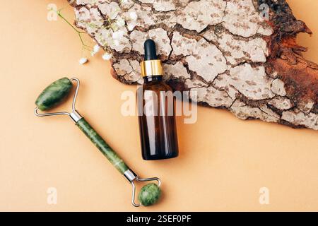 Jade facial roller and amber glass dropper bottle on a beige background with tree bark and flowers. Skin care cosmetic concept. Top view, flat lay. Stock Photo